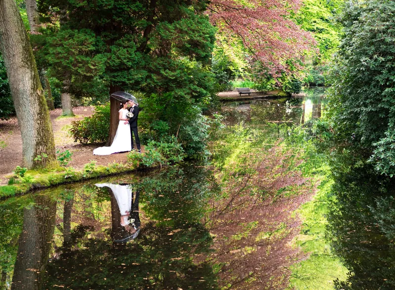 Bruidspaar onder een paraplu aan het water, gespiegeld in een vijver omringd door groen