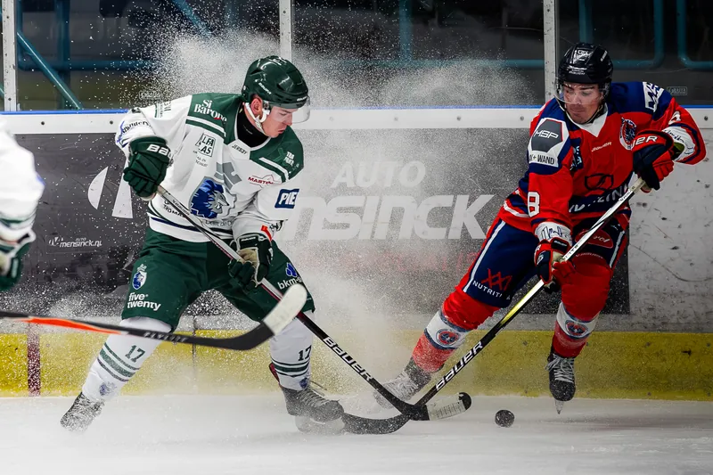 Twee ijshockeyers vechten om de puck langs de boarding terwijl het ijs opspat