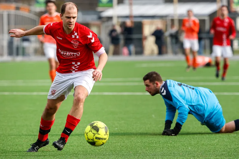Voetballer in rood shirt dribbelt weg bij een tegenstander op de grond