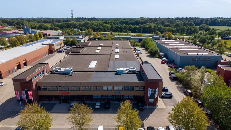 Dronefoto van een grote bedrijfshal met kantoorvleugel en bomen langs de gevel