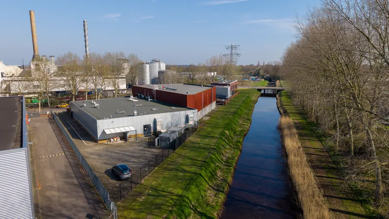 Dronefoto van een bedrijfshal naast een kanaal met industrieel terrein op de achtergrond